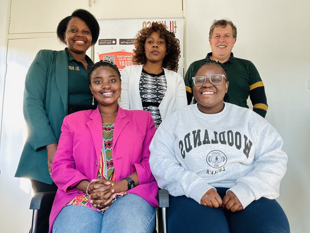 5 people smiling, one man is standing on the right next to two women, while two women are sitting